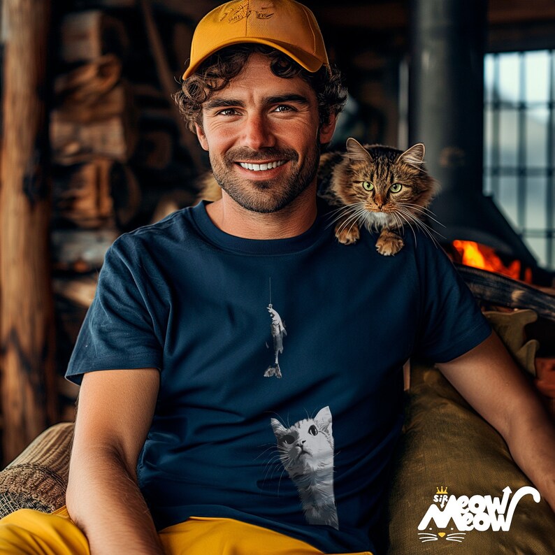 Fisherman sitting on a cozy chair with his cat perched on his shoulder. He is wearing a navy tshirt with the illustration of a cat starring up at a fish dangling from a fish line.