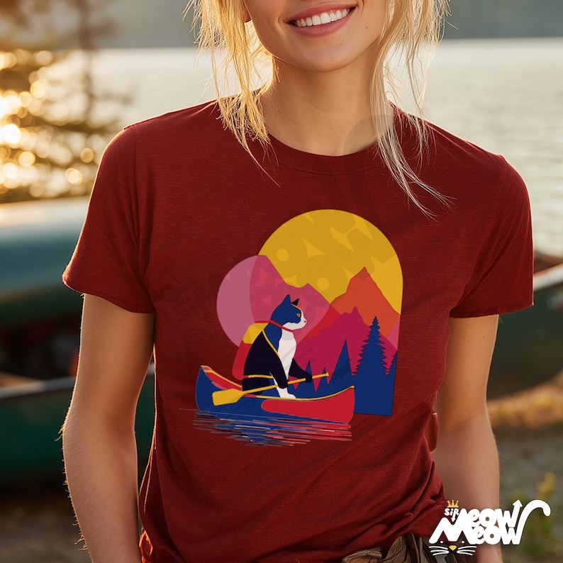 Puede incluir: Una camiseta granate con un gr&aacute;fico de un gato en una canoa en un lago, con un fondo de monta&ntilde;a y puesta de sol. El gato lleva una mochila y rema. La camiseta tiene el texto "Sir Meow Meow" en la parte inferior.