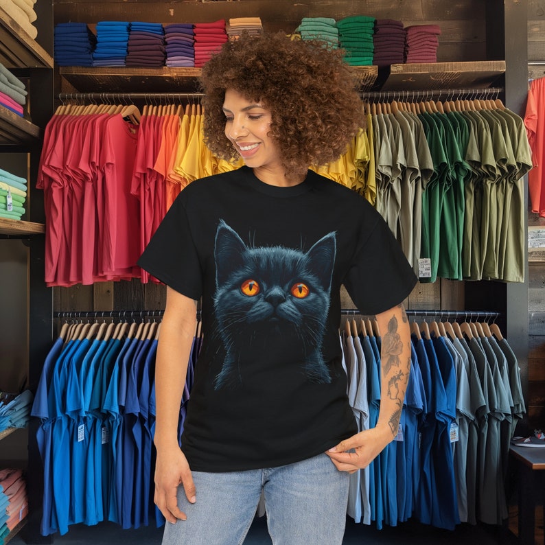 May include: Black t-shirt featuring a detailed illustration of a black cat with bright orange eyes. The cat's fur is depicted with fine detail, creating a striking contrast against the dark background. The t-shirt is a classic crew neck style.