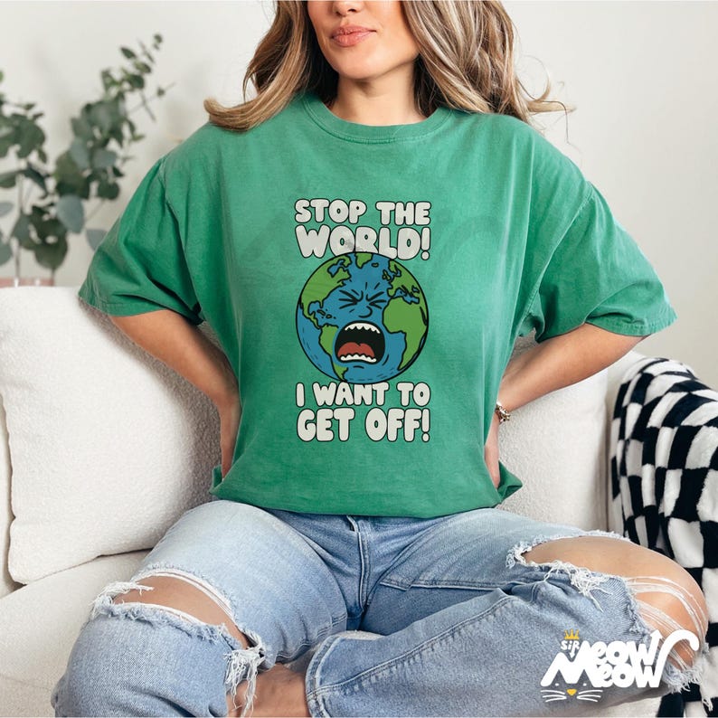 Może przedstawiać: Zielona koszulka z napisem "STOP THE WORLD! I WANT TO GET OFF!" nad grafiką Ziemi w opałach. Koszulka jest w jednolitym zielonym kolorze i ma kr&oacute;tkie rękawy. Modelka ma na sobie podarte niebieskie dżinsy.