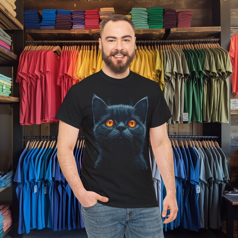 May include: Black t-shirt featuring a detailed illustration of a black cat with bright orange eyes. The cat's fur is depicted with intricate shading, creating a realistic appearance. The t-shirt is made of a soft material and is suitable for casual wear.