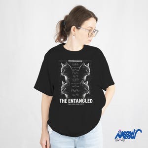 May include: Black t-shirt featuring a graphic design with four cat portraits and scientific diagrams. The text "THE ENTANGLED TWO CATS, ONE FATE" is printed below the images. The shirt is worn by a person.