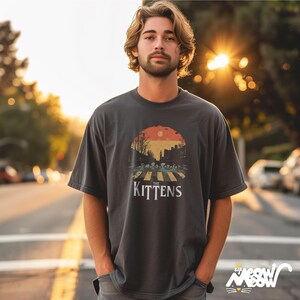 May include: A dark gray t-shirt with a vintage-style graphic of kittens crossing a street. The graphic features a sunset, trees, and the text "The Kittens".