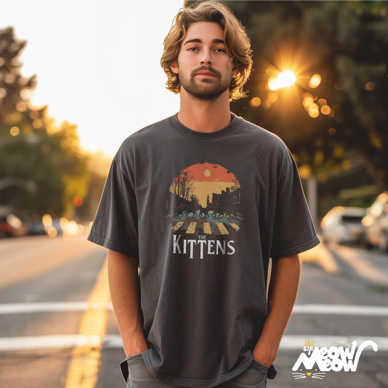 May include: A dark gray t-shirt with a vintage-style graphic of kittens crossing a street. The graphic features a sunset, trees, and the text "The Kittens".