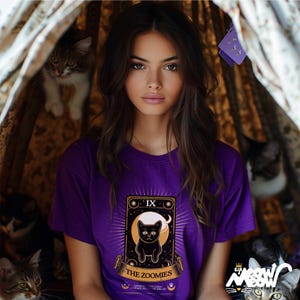 Puede incluir: Una mujer con una camiseta morada con una ilustraci&oacute;n de un gato negro y el texto "The Zoomies" en un dise&ntilde;o de carta de tarot. La carta est&aacute; rodeada de estrellas y una luna creciente.