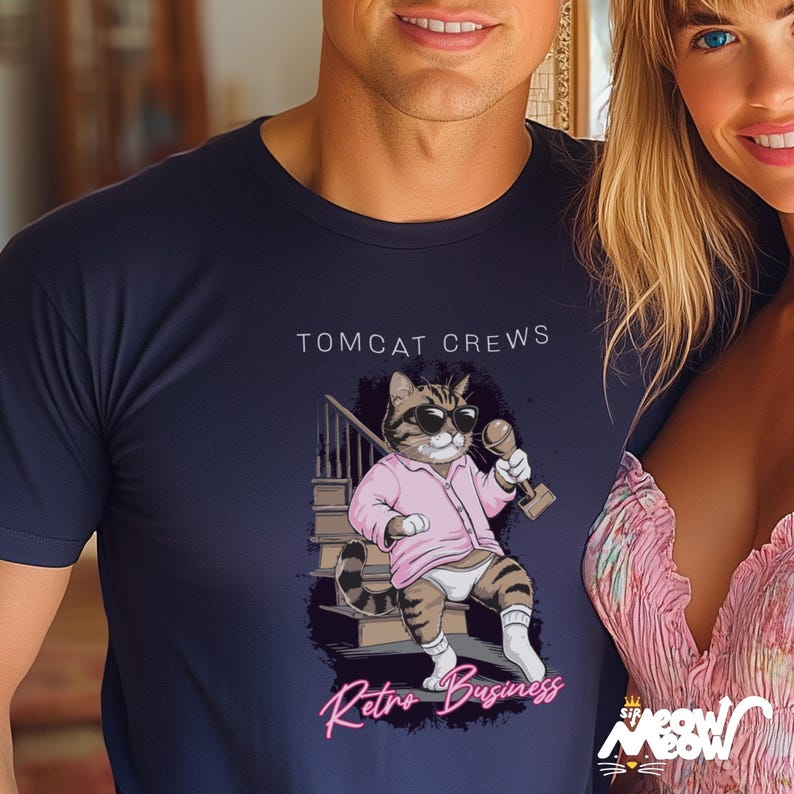 May include: A navy blue t-shirt with a cartoon cat wearing a pink shirt and sunglasses holding a microphone. The text "Tomcat Crews Retro Business" is printed on the shirt.