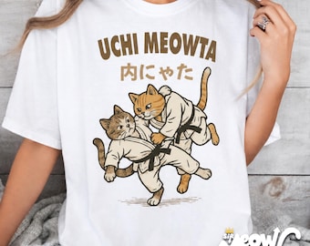 Puede incluir: Una camiseta blanca con un gráfico de dos gatos vestidos con uniformes de artes marciales blancos y cinturones negros, realizando un movimiento de judo. El texto UCHI MEOWTA y caracteres japoneses están impresos en marrón sobre los gatos.