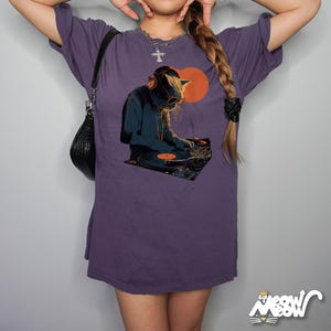K&ouml;nnte beinhalten: Lilafarbenes T-Shirt mit einem Aufdruck einer Katze als DJ mit Kopfh&ouml;rern und Sonnenbrille, die Schallplatten auflegt. Das Design beinhaltet eine orangefarbene Sonne und einen Plattenspieler. Das Shirt hat eine lockere Passform.
