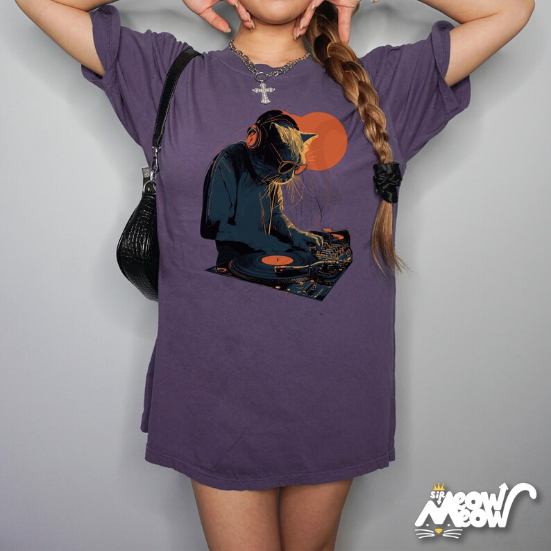 K&ouml;nnte beinhalten: Lilafarbenes T-Shirt mit einem Aufdruck einer Katze als DJ mit Kopfh&ouml;rern und Sonnenbrille, die Schallplatten auflegt. Das Design beinhaltet eine orangefarbene Sonne und einen Plattenspieler. Das Shirt hat eine lockere Passform.
