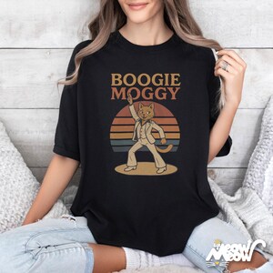 May include: Black t-shirt with the text "BOOGIE MOGGY" above a cartoon cat in a white disco suit, set against a retro sunset design. The cat is raising one arm in a dance pose. The shirt is a casual wear item.