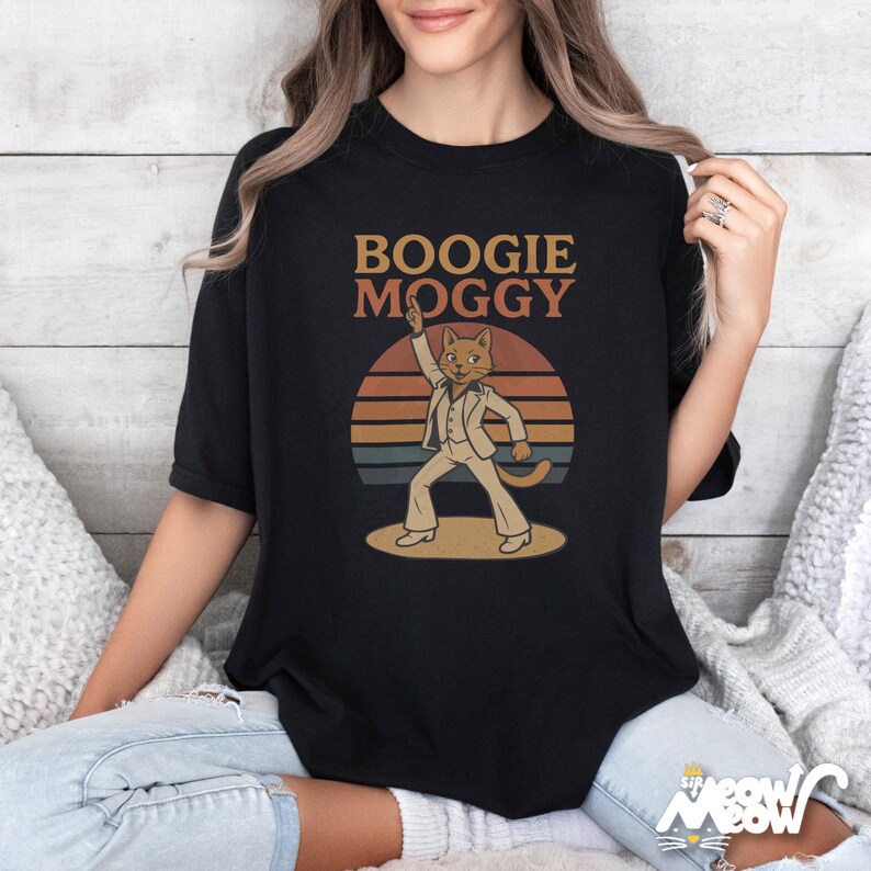 May include: Black t-shirt with the text "BOOGIE MOGGY" above a cartoon cat in a white disco suit, set against a retro sunset design. The cat is raising one arm in a dance pose. The shirt is a casual wear item.