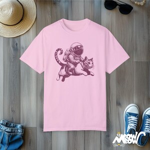 Bloom pink colored Comfort Colors 1717 unisex T-shirt with a humorous white graphic of an astronaut riding atop a playful cat, combining outer space and cat lover themes.