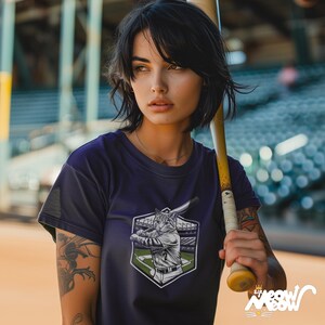 May include: A woman wearing a purple t-shirt with a graphic of a cat playing baseball. The cat is wearing a baseball uniform and holding a bat. The woman is holding a baseball bat and looking to the side.