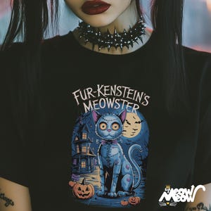 Może przedstawiać: Czarny t-shirt z ilustracją kota z kresk&oacute;wki i napisem "Fur-kenstein's Meowster". Projekt zawiera upiornego kota, dynie, nawiedzony dom i pełnię księżyca, idealny na Halloween.