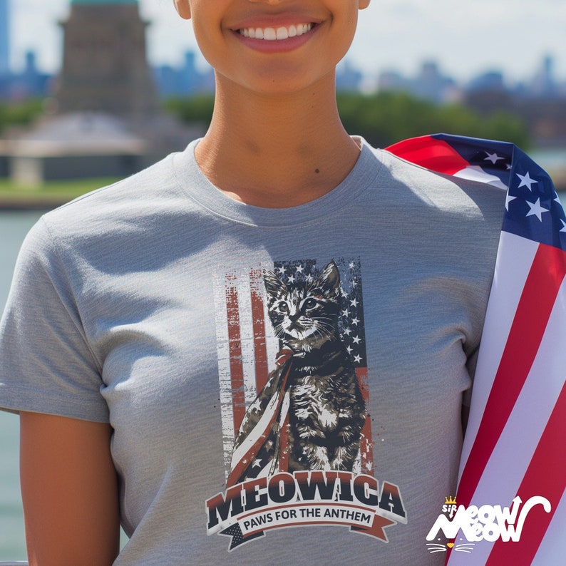May include: A gray t-shirt with a graphic of a cat wearing a red, white, and blue American flag cape. The text "MEOWICA PAWS FOR THE ANTHEM" is printed below the cat.