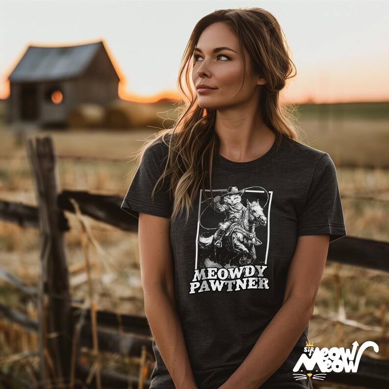 Pode incluir: Uma mulher vestindo uma camiseta cinza escura com uma ilustra&ccedil;&atilde;o em preto e branco de um gato montando um cavalo. O texto "MEOWDY PAWTNER" est&aacute; impresso abaixo da ilustra&ccedil;&atilde;o.