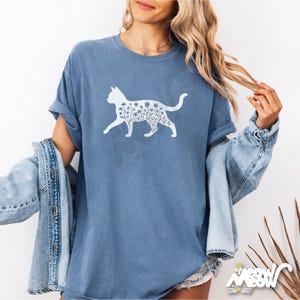 May include: A blue t-shirt featuring a white cat silhouette filled with floral designs. The shirt has short sleeves and a relaxed fit. The image also shows a denim jacket and the text "Meow Meow" in the bottom right corner.