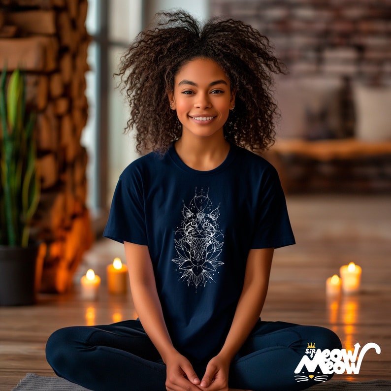 May include: Navy blue t-shirt with a white graphic of a cat design. The graphic includes a cat's face, geometric shapes, and a lotus flower. The shirt has short sleeves and a crew neck. The text "Sip Meow Meow" is at the bottom right.