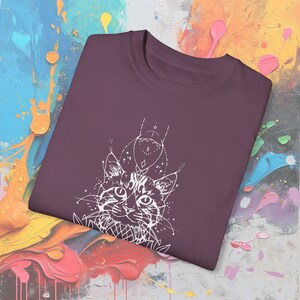 May include: A maroon crew neck t-shirt featuring a white graphic of a cat's face with geometric and floral accents. The cat's eyes are detailed, and the design has a bohemian aesthetic. The shirt is made of a soft material.