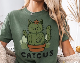 Può includere: Una maglietta verde con un design di cactus gatto. Il cactus è un cactus verde con un fiore in cima e un viso sorridente. Il testo "CATCUS" è stampato sotto il cactus.