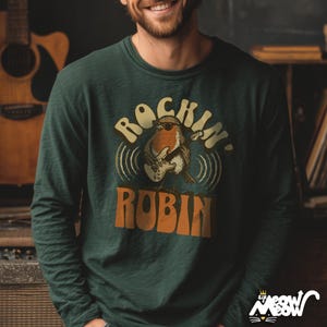 Könnte beinhalten: Dunkelgrünes Langarmshirt mit dem Text "Rockin' Robin" und einem Cartoon-Rotkehlchen, das E-Gitarre spielt. Das Rotkehlchen trägt eine Sonnenbrille. Das Shirt hat ein Retro-Design.