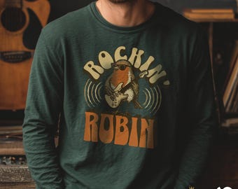 Puede incluir: Camiseta de manga larga verde oscuro con el texto "Rockin' Robin" y un petirrojo de dibujos animados tocando la guitarra eléctrica. El petirrojo lleva gafas de sol. Diseño retro.