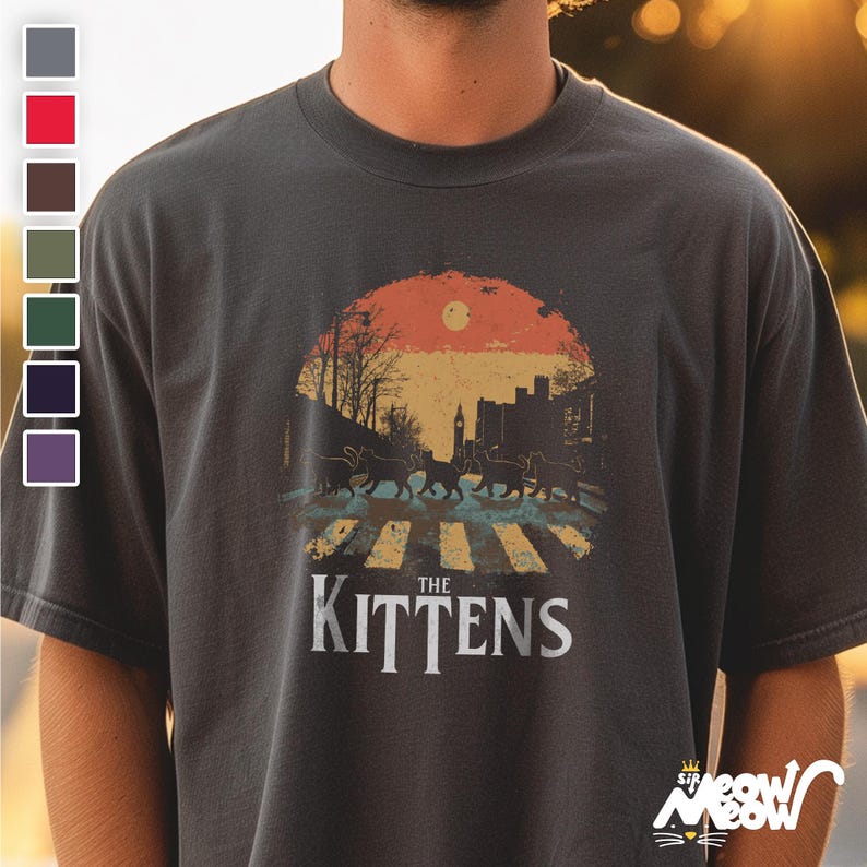 May include: A dark gray t-shirt with a retro-style graphic of a street scene with five black cats walking across a crosswalk. The graphic includes a sunset, a building, and the text "The Kittens".
