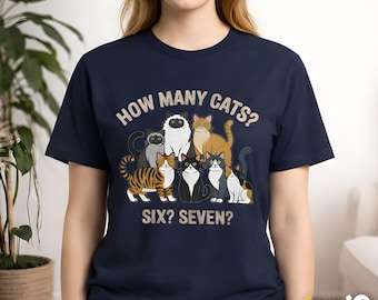 Puede incluir: Camiseta azul marino con un gráfico de siete gatos de dibujos animados y el texto "HOW MANY CATS? SIX? SEVEN?". Los gatos son de varios colores y patrones. La camiseta es de cuello redondo.