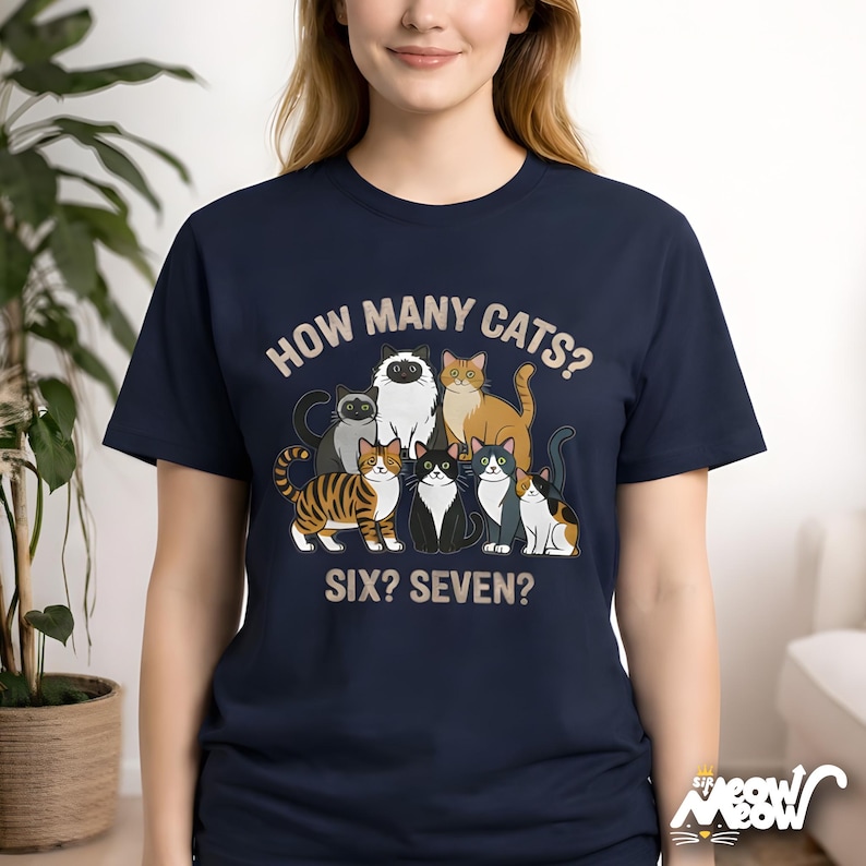 May include: Navy blue t-shirt with a graphic of seven cartoon cats and the text "HOW MANY CATS? SIX? SEVEN?". The cats are in various colors and patterns. The shirt is a crew neck.
