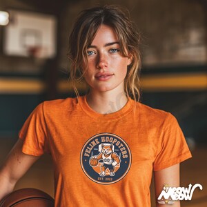 K&ouml;nnte beinhalten: Orangefarbenes T-Shirt mit einem kreisf&ouml;rmigen Aufdruck, der einen Cartoon-Fuchs beim Basketballspielen zeigt, sowie dem Text "Feline Hoopsters" und "Est. 2023". Das T-Shirt wird von einer Person getragen, die einen Basketball in einer Turnhalle h&auml;lt.