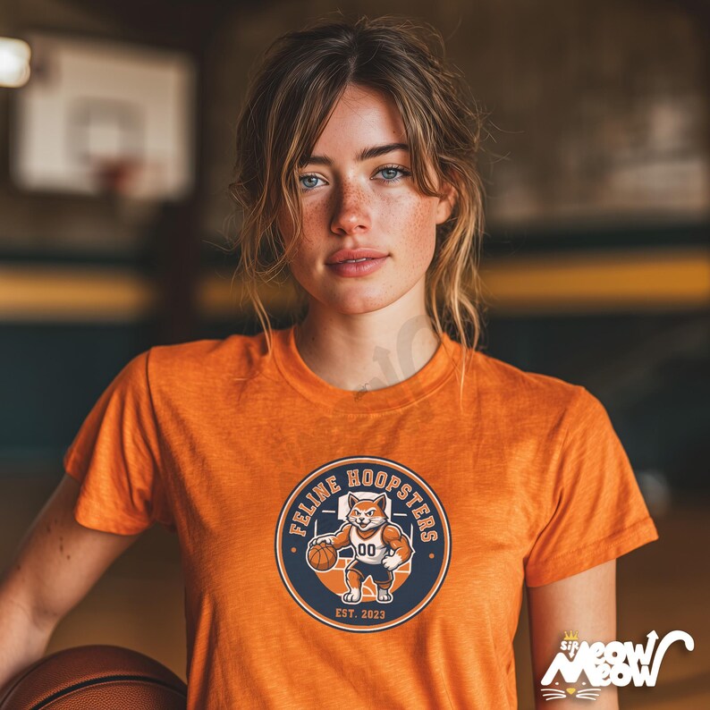 K&ouml;nnte beinhalten: Orangefarbenes T-Shirt mit einem kreisf&ouml;rmigen Aufdruck, der einen Cartoon-Fuchs beim Basketballspielen zeigt, sowie dem Text "Feline Hoopsters" und "Est. 2023". Das T-Shirt wird von einer Person getragen, die einen Basketball in einer Turnhalle h&auml;lt.