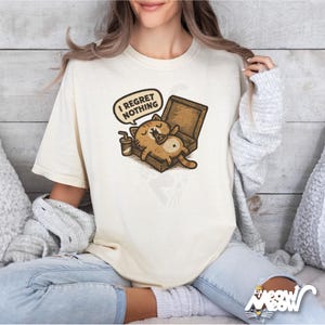 K&ouml;nnte beinhalten: Cremefarbenes T-Shirt mit einer Cartoon-Katze, die sich in einer Pizzaschachtel entspannt, mit dem Text "I Regret Nothing" in einer Sprechblase. Ein kleiner Kaffeebecher steht daneben. Das Shirt hat eine lockere Passform.
