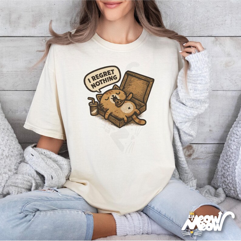 K&ouml;nnte beinhalten: Cremefarbenes T-Shirt mit einer Cartoon-Katze, die sich in einer Pizzaschachtel entspannt, mit dem Text "I Regret Nothing" in einer Sprechblase. Ein kleiner Kaffeebecher steht daneben. Das Shirt hat eine lockere Passform.