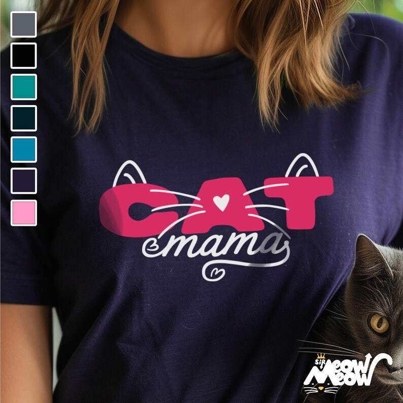 May include: Navy blue t-shirt with a pink "CAT" design, featuring cat ears and a heart, and the word "mamas" in white script. A gray cat is visible on the right side of the image. The shirt has a crew neck.