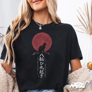 Op de afbeelding: Zwart T-shirt met een katten silhouet tegen een rode zon, en Japanse tekst. Het ontwerp bevat de zinnen "Stand Up 9 Times" en "Fall Down 8 Times". Het shirt heeft een relaxte pasvorm.