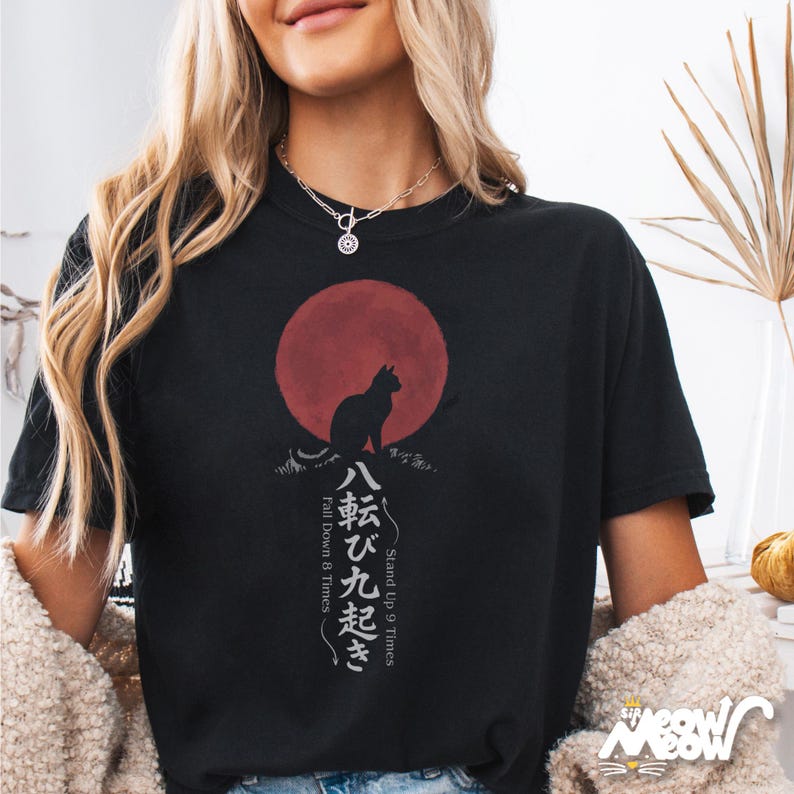 Op de afbeelding: Zwart T-shirt met een katten silhouet tegen een rode zon, en Japanse tekst. Het ontwerp bevat de zinnen "Stand Up 9 Times" en "Fall Down 8 Times". Het shirt heeft een relaxte pasvorm.