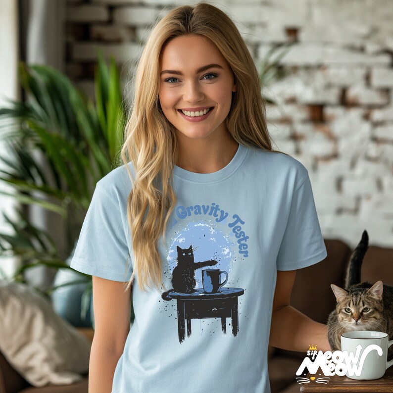 May include: A light blue t-shirt featuring a black cat graphic and the text "Gravity Tester". The cat is reaching for a cup of coffee on a table.