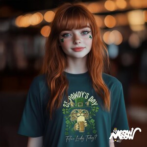 May include: A teal green t-shirt featuring a graphic of a cat wearing a green leprechaun hat and holding a pint of beer. The text on the shirt reads "St. Pawdy's Day" and "Feline Lucky Today?"