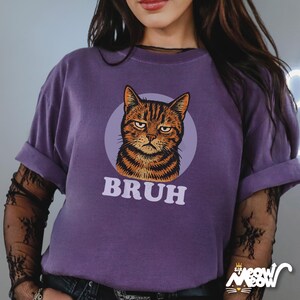 May include: A purple t-shirt with a grumpy cat illustration in a light purple circle, and the word "BRUH" underneath. The cat is orange and brown striped. The sleeves are rolled up.