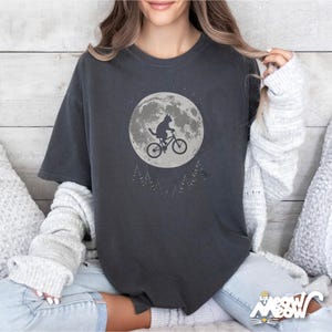 May include: Dark gray t-shirt featuring a cat riding a bicycle in front of a full moon. The design includes a silhouette of a cat on a bike, a large moon, and a few trees. The text "Sip Meow Meow" is at the bottom right.