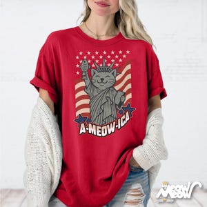 May include: Red t-shirt featuring a cartoon cat as the Statue of Liberty, holding a fish and a book, with the text "A-MEOW-ICA" and a flag design. The shirt has a relaxed fit.