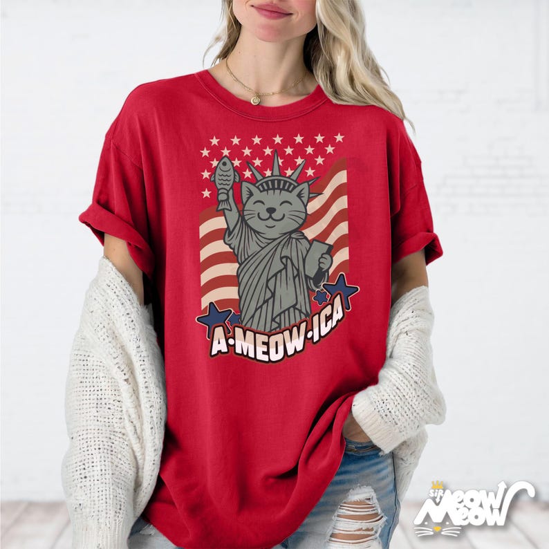 May include: Red t-shirt featuring a cartoon cat as the Statue of Liberty, holding a fish and a book, with the text "A-MEOW-ICA" and a flag design. The shirt has a relaxed fit.