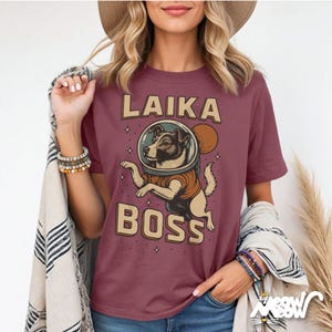 K&ouml;nnte beinhalten: Burgunderfarbenes T-Shirt mit einem Retro-Design, das einen Hund in einem Weltraumhelm und die Worte "LAIKA BOSS" zeigt. Die Hundeillustration ist in Braun-, Beige- und Schwarzt&ouml;nen gehalten, mit einem Planeten und Sternen. Ein lustiges, Weltraum-Themen-Grafik-T-Shirt.