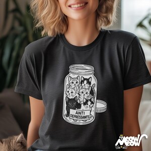 May include: Black t-shirt featuring a jar filled with several cartoon cats. The jar has a white label that reads "ANTI DEPRESSANTS". The shirt has a round neck and short sleeves. The image is a playful design.