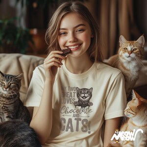 May include: A woman wearing a cream colored t-shirt with a graphic of a cat and the text "My two great loves Chocolate and Cats!" She is holding a chocolate bar in her hand and smiling at the camera. There are three cats in the image, one sitting on the couch behind her and two sitting in front of her.