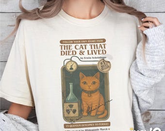 May include: A white t-shirt with a vintage-style illustration of a cat in a box. The text reads "The Cat That Died & Lived" by Erwin Schrödinger. The illustration includes a beaker, a radiation symbol, and a signature. The text "#1 Best Seller" is also included.