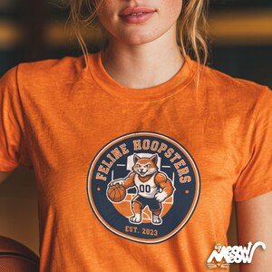 K&ouml;nnte beinhalten: Orangefarbenes T-Shirt mit einem Cartoon-Katzenmotiv, das Basketball spielt. Das Design zeigt die Worte "Feline Hoopsters" in einem kreisf&ouml;rmigen marineblauen Emblem mit "EST. 2023" darunter. Die Katze tr&auml;gt ein Trikot mit der Nummer "00".