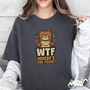 Op de afbeelding: Een donkergrijs T-shirt met een cartoon oranje kat die een vork en mes vasthoudt. De tekst "WTF WHERE'S THE FOOD?" is onder de kat gedrukt.