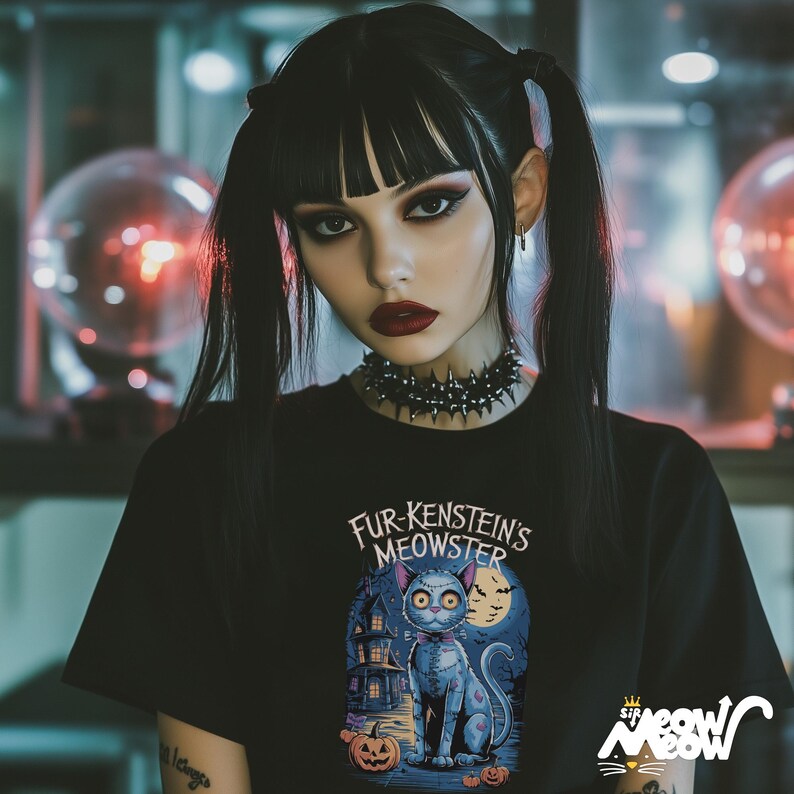 Może przedstawiać: Czarny t-shirt z napisem "Fur-Kenstein's Meowster" i ilustracją kota z kresk&oacute;wki. Kot jest niebieski z muszką, przed nawiedzonym domem i dyniami. Koszulka ma r&oacute;wnież logo "sip meow meow".