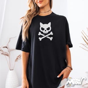 May include: Black t-shirt featuring a white graphic of a skull with cat ears and crossed bones. The shirt has short sleeves and a relaxed fit. The design is centered on the chest.